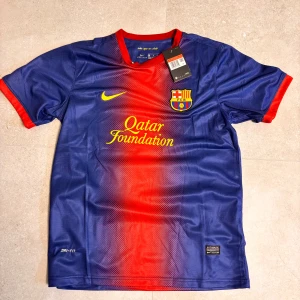 FC Barcelona matchtröja  - Säljer en officiell FC Barcelona fotbollströja från Nike med Messi 10 på ryggen. Tröjan är blå med röd mitt, gula detaljer och klubbmärke på bröstet. Tillverkad i lätt och ventilerande Dri-FIT-material, perfekt för fotboll eller att bära som supporter.