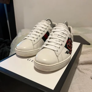 Gucci vita sneakers med broderad orm - Säljer ett par vita Gucci sneakers med klassiska gröna och röda ränder på sidan samt en broderad orm. Baksidan har metallicröd och metallicgrön detalj med Gucci-logga. Skorna har snörning och platt sula.