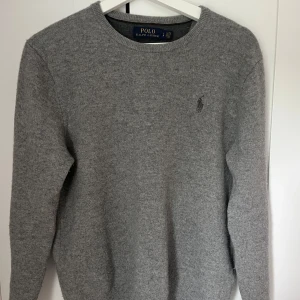 Grå stickad tröja från Polo Ralph Lauren - Klassisk grå stickad tröja från Polo Ralph Lauren med rund halsringning och diskret broderad logga på bröstet. Perfekt basic-plagg med lång ärm och mjuk känsla, passar till allt och är superenkel att styla.