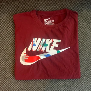 Vinröd Nike t-shirt med färgglad logga - Snygg vinröd t-shirt från Nike i bomull med klassisk passform. Stor Nike-logga framtill med färgglada detaljer och mindre loggor på ärm och rygg. Perfekt för dig som gillar streetwear och sportig stil. Det står att det ska vara storlek L men det är mer storlek S