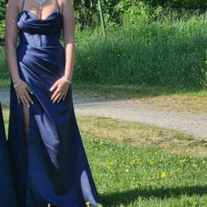 Mörkblå långklänning med snörning i ryggen - Säljer en elegant mörkblå långklänning med smala axelband och draperad byst från jj house. Klänningen har en snygg slits framtill och snörning i ryggen för perfekt passform (obs, snöret är lite långt men går att förkorta) Tillverkad i ett glansigt material som ger en lyxig känsla. Har använt en gång och har kvar orginal påsen att förvara den i. Denna klänning är perfekt för dig som ska gå på bal eller fest💕