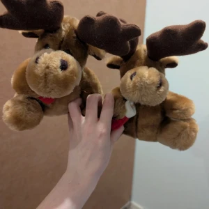 Gosedjur i form av älgar - Bilderna visar två mjukisdjur i form av älgar, inte en bok. De är bruna med stora horn och röda detaljer. Perfekta som gosedjur eller present till någon som gillar djur.