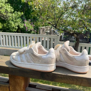 Beige Adidas sneakers med breda snören - Säljer ett par beige sneakers från Adidas med breda, platta snören och klassiska tre ränder på sidan. Skorna har en vit sula och detaljer i mocka och syntet. Skorna är använda och har några enstaka skador inuti skon men funkar precis som nya ändå. Lite smutsiga också men går att tvätta! 