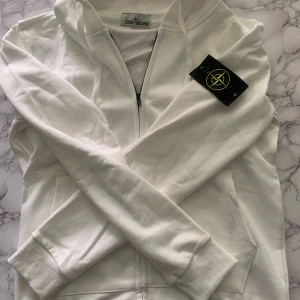 Vit hoodie med dragkedja från Stone Island - Säljer en stilren vit hoodie från Stone Island med dragkedja framtill och klassisk logotyp-patch på ärmen. Tröjan har huva med dragsko och två fickor framtill. Perfekt för en clean och avslappnad look.