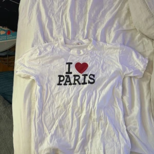 Vit t-shirt med I ❤️ PARIS-tryck - Vit t-shirt från Alam Fashion Premium med det klassiska trycket 'I ❤️ PARIS' på bröstet. T-shirten har rund halsringning och är tillverkad i 100% bomull, vilket gör den mjuk och skön att bära. Pris går alltid att diskutera❣️