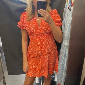 Röd blommig kortklänning med volang - Säljer en söt röd kortklänning med vita blommor och volangdetaljer på axlarna. Klänningen har v-ringning och korta puffärmar. As fin!!!