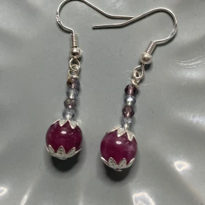 Handgjorda örhängen med lila pärlor - Säljer ett par unika handgjorda örhängen med stora lila turmalin pärlor och mindre glittriga kristaller i silver och lila nyanser. Örhängena har silverfärgade detaljer och krokar. Perfekta för att ge din stil en färgklick!