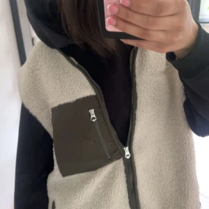 Beige fleeceväst med grön ficka - Säljer en fleeceväst med dragkedja framtill och en grön bröstficka med dragkedja. Perfekt att slänga över en hoodie för extra värme. Västen avslappnad look. Storlek L men passar mig som vanligtvis har S/M