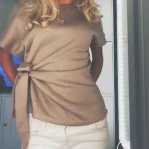 Beige topp med knyt i sidan från Zara - Säljer en beige topp från Zara med korta ärmar och snygg knytning i sidan. Toppen har rund halsringning och är tillverkad i ett mjukt material som ger en avslappnad men stilren look.