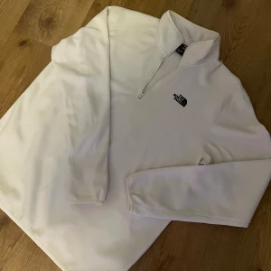 Vit fleece-tröja från The North Face - Säljer en vit fleece-tröja från The North Face med halv dragkedja och broderad logga på bröstet. Perfekt för lager-på-lager och håller dig varm under kyliga dagar. Klassisk och stilren design med lång ärm.