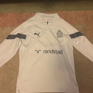 Olympique de Marseille fotbollströja  - Säljer en vit långärmad fotbollströja från Puma för Olympique de Marseille. Tröjan har halv dragkedja, klubbmärke och Puma-logga på bröstet samt sponsortryck från Randstad. Tillverkad i ett lätt och sportigt material, perfekt för träning eller match. Pris kan diskuteras!! Tveka inte att skriva!!