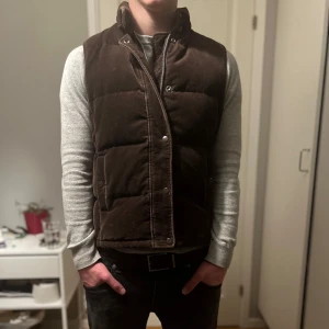 Brun manchester väst - Säljer en brun dunväst i manchester från GAP med tryckknappar framtill. Västen har quiltad design och två fickor på sidorna. Perfekt att slänga över en hoodie eller tröja för extra värme och stilpoäng. 