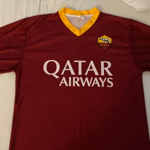 AS Roma matchtröja vinröd Qatar Airways - Snygg vinröd AS Roma fotbollströja med gult V-hals och klubbmärke på bröstet. Qatar Airways-logga tryckt på framsidan. Tillverkad i lätt och ventilerande material, perfekt för match eller träning.