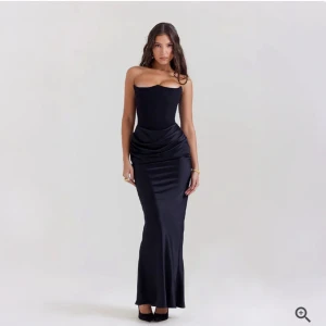 Svart långklänning med draperad midja - Elegant svart långklänning med axelbandslös design och draperad detalj vid midjan. Klänningen har en figurnära passform och är tillverkad i ett glansigt material som ger en lyxig känsla.