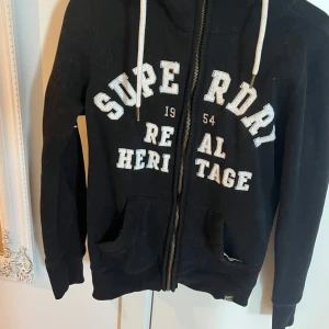 Svart hoodie från Superdry med dragkedja - Svart hoodie från Superdry med vit broderad logga på bröstet och ärmen. Jackan har huva med dragsko, två fickor fram och dragkedja. Insidan är mjuk och mysig, perfekt för kyliga dagar.