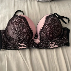 Rosa och svart spets-bh med detaljer - Säljer en push-up bh i ljusrosa med svart spets och dekorativa banddetaljer framtill. Den är använd endast 1 gång. Bh:n har vadderade kupor, justerbara axelband och hakknäppning bak. Spetsen har blommigt mönster och kanten är vågig för en extra snygg look. Finns matchande trosor om man vill ha. 