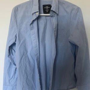 Ljusblå skjorta från L.O.G.G. H&M - Klassisk ljusblå skjorta med diskreta ränder från L.O.G.G. H&M. Skjortan har lång ärm, bröstficka och knäppning framtill. Den är i storlek 