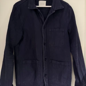 Mörkblå overshirt från A Day's March - Stilren mörkblå overshirt från A Day's March med tre stora fickor framtill och klassisk krage. Jackan är tillverkad i ett kraftigare bomullstyg och har knäppning med svarta knappar. Perfekt att slänga över en hoodie eller t-shirt.