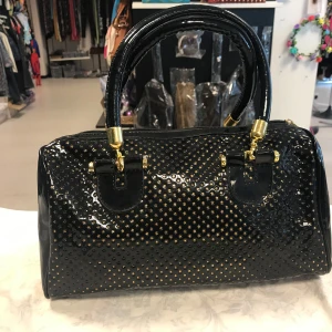 Svart lackad handväska med gulddetaljer - Snygg svart handväska i glansigt lackmaterial med små guldfärgade nitar på framsidan. Väskan har två stadiga handtag och guldfärgade metalldetaljer, samt dragkedja upptill. Perfekt för dig som vill sticka ut med en elegant accessoar. Helt ny 