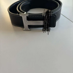 Svart skärp med silverfärgat H-spänne - Säljer ett svart bälte med tydliga vita sömmar och ett stort H-format spänne i silverfärg. Bältet är gjort i skinn och har en klassisk, stilren design som passar till många olika outfits.