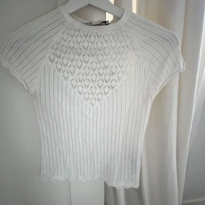 Vit stickad topp med spetsdetaljer - Säljer en vit stickad topp med korta ärmar och vackert spetsmönster framtill. Toppen har rund halsringning och ett diskret vågigt avslut nertill. Perfekt för en fräsch och stilren look.