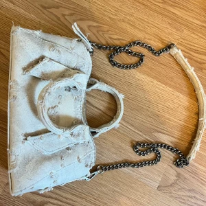 Beige jeansväska med kedja - Cool beige handväska i jeansmaterial med slitna detaljer och silverfärgad kedja som axelrem. Väskan har dubbla handtag, dragkedja upptill💕