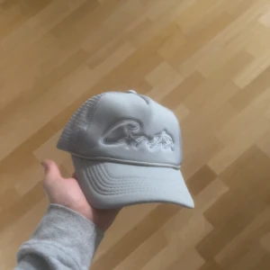 Ljusgrå keps med broderad text från corteiz - Snygg ljusgrå keps med broderad text framtill. Kepsen har böjd skärm och justerbar snapback baktill i meshmaterial för extra ventilation. Perfekt för en stilren look