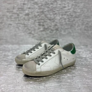 Vita sneakers med grå snörning och gröna detaljer från Golden Goose - Säljer ett par vita sneakers från Golden Goose med grå snörning, grå sula och gröna detaljer på hälen. Skorna har perforerade stjärndetaljer på sidan och klassisk låg modell. Perfekta för en avslappnad och trendig stil. De flesta storlekar finns tillgängliga. Leverans sker inom 12–15 dagar.