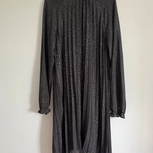 Svart knälångklänning med prickigt mönster från Orsay - Säljer en svart knälångklänning från Orsay med silver/guld prickigt mönster. Klänningen har långa ärmar med resår och volang vid ärmslut samt knytband i halsen. Kan välja att knyta fram eller bak. Tunt och lätt material som faller snyggt. Fodrad inuti. Funkar bra för XS-S.