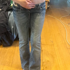 Lågmidjade bootcut jeans från True Religion - Säljer ett par blå lågmidjade jeans från True Religion med bootcut passform. Jeansen har slitna detaljer, kontrastsömmar och dekorativa fickor med knapp och broderi bak. Perfekta för dig som gillar en klassisk och cool jeansstil.