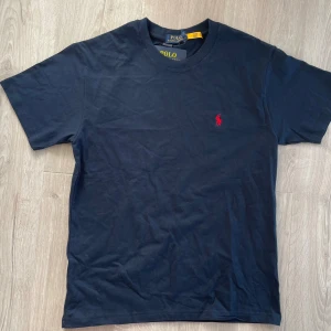 Marinblå t-shirt från Polo Ralph Lauren - Säljer en klassisk mörkblå t-shirt från Polo Ralph Lauren med gul broderad logga på bröstet. T-shirten har rund hals och korta ärmar. Perfekt basplagg med stilren design.