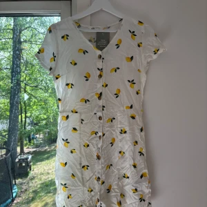 Vit klänning med citronmönster från H&M - Söt vit klänning från H&M med korta ärmar och knappar framtill. Klänningen har citronmönster i gult och grönt, perfekt för varma dagar. Den är i barnstorlek 164 som passar XXS / XS / S