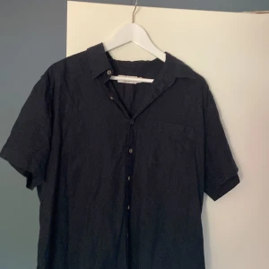 Svart linneskjorta från Dressmann - Svart kortärmad skjorta från Dressmann i linnemix. Klassisk krage, knäppning framtill och bröstficka. Perfekt för dig som gillar en clean och stilren look med lite loose fit.