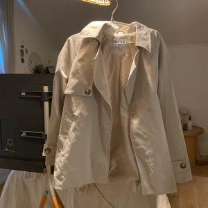 Beige trenchcoat från NA-KD med bälte - Snygg beige trenchcoat från NA-KD med klassisk krage, knappar och bälte i midjan. Jackan har långa ärmar med knäppning vid ärmslut och är tillverkad i ett mjukt bomullsmaterial. Perfekt för vår och höst när du vill ha en clean och stilren look.