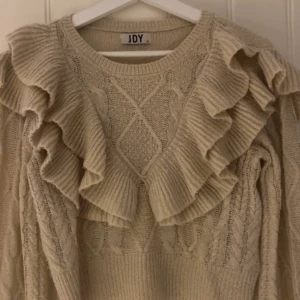 Beige stickad tröja med volang från JDY - Superfin beige stickad tröja från JDY med stora volanger framtill och längs axlarna. Tröjan har ett snyggt stickat mönster och rund halsringning. Perfekt för dig som gillar detaljer och vill ha något extra i garderoben.