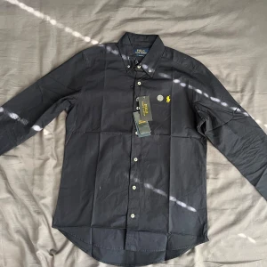 Helt nyt blå skjorta från Polo Ralph Lauren - Säljer en mörk blå långärmad skjorta från Polo Ralph Lauren i slim fit-modell. Skjortan har klassisk krage, knappar framtill och den ikoniska gula broderade loggan på bröstet. Perfekt för dig som gillar stilrena plagg med snygga detaljer.