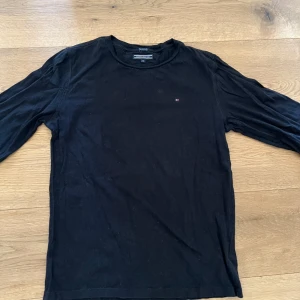 Svart långärmad tröja från Tommy Hilfiger - Säljer en svart långärmad tröja från Tommy Hilfiger i mjuk bomull. Tröjan har en rund halsringning och en liten broderad logga på bröstet. Perfekt basplagg med stilren design. Passar XS troligen