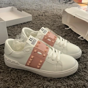 Valentino Garavani sneakers med nitar - Säljer ett par vita Valentino Garavani sneakers med ljusrosa  nitar över vristen. Skorna har rund tå, platt sula och klassisk snörning. Perfekta för dig som vill ha en lyxig och trendig look.