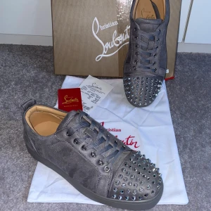 Grå sneakers med nitar från Christian Louboutin - Säljer ett par gråa sneakers från Christian Louboutin med silverfärgade nitar på tån och klassisk röd sula. Skorna har snörning och är tillverkade i mocka med en rund tå. Perfekta för dig som vill sticka ut med din stil.                             - helt nya och oanvända. Storlek 40 men passar upp till 43 eftersom att de inte är formade än