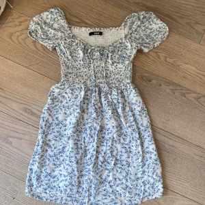 Blommig kortklänning från BikBok - Supersöt vit kortklänning med blått blommönster från BikBok. Klänningen har puffärmar, smock över byst och midja samt knyt framtill. Perfekt för sommarfesten!