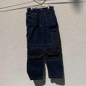 Mörkblå cargopants från PX - Mörkblå cargopants från PX med kontrasterande svarta fickor och vita sömmar. Byxorna har flera stora fickor framtill och på sidorna, samt dragkedja och knapp i midjan. Tillverkade i 100% bomull, storlek 46.