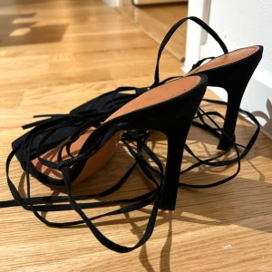 Svarta klackskor med snörning - Säljer ett par svarta klackskor med höga klackar och långa tunna snören som lindas runt benet. Skorna har fyrkantig tå och är i mockaliknande material. Perfekta för att lyfta en outfit med trendig och elegant känsla.