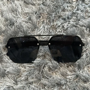 Svarta solglasögon med geometrisk form - Otroligt robusta!! Snygga svarta solglasögon med unika, kantiga linser och metallbåge. Glasögonen har en modern och edgy design med mörka linser och silverdetaljer vid skruvarna. Perfekta för dig som vill sticka ut med en cool accessoar. 