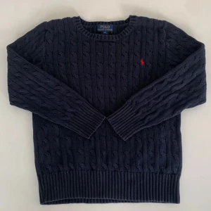 Mörkblå kabelstickad tröja Polo Ralph Lauren - Kabelstickad mörkblå tröja från Polo Ralph Lauren med rund halsringning och röd broderad logga på bröstet. Tröjan har långa ärmar och ribbade muddar vid ärmslut och nederkant. Perfekt för dig som gillar klassisk och stilren look.