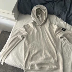 Beige hoodie från Calvin Klein - Snygg beige hoodie från Calvin Klein med klassisk huva och stor magficka. Tröjan har lång ärm och en svart CK-logga på vänster ärm.  Den är använd ett fåtal gånger och har en liten fläck på sig som ni kan se i sista bilden. Fläcken är knappt märkbar och går säkert bort i tvätten 