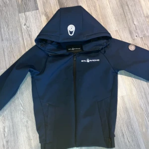 Marinblå jacka med huva från Sail Racing - Säljer en marinblå jacka från Sail Racing med huva och dragkedja framtill. Jackan har logga på bröstet och på huvan, samt två sidofickor. Materialet är slätt och jackan är fodrad med fleece på insidan för extra komfort.
