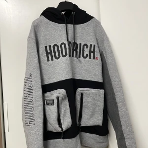 Grå och svart hoodie från Hoodrich - Säljer en snygg grå och svart hoodie från Hoodrich i storlek L. Den har en stor logga på bröstet och ärmarna, samt praktiska fickor med dragkedja framtill. Perfekt för streetwear-stilen och håller dig varm och bekväm. Hör av dig om du är intresserad!