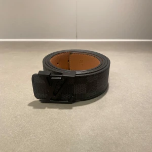 Svart LV bälte - Snyggt svart bälte från Louis Vuitton med klassiskt Damier-rutmönster och mattsvart LV-spänne. Bältet är gjort i exklusivt skinn med brun insida och har en bred, stilren design som verkligen sticker ut. Perfekt accessoar för att lyfta din outfit.