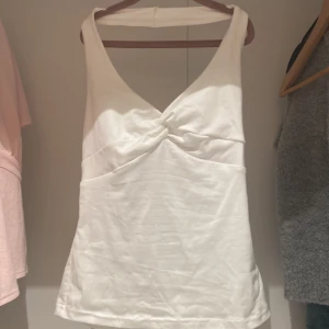 Vit ärmlös halterneck linnetopp - Snygg vit linnetopp med halterneck och twistad detalj framtill. Toppen är ärmlös och har en djup v-ringning som ger en fräsch och modern look. Perfekt att matcha med jeans eller kjol för en trendig stil.