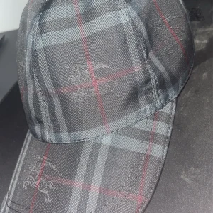 Rutig keps från Burberry i svart och grått - Snygg keps från Burberry med klassiskt rutigt mönster i svart, grått och röda linjer. Kepsen har broderade Burberry-loggor och justerbart spänne i svart skinn baktill. Perfekt accessoar för en trendig look.